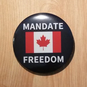 Può includere: Un bottone nero e bianco con il testo "MANDATE FREEDOM" e una bandiera canadese al centro.
