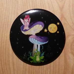 May include: A circular black button with a fantasy illustration. A fairy sits on a purple and white mushroom, with a frog below. The background depicts a starry night with a smiling moon and stars. A whimsical design.