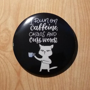 May include: A black and white button featuring a white cat holding a cup of coffee. The text on the button reads "I run on caffeine, chaos and cuss words".
