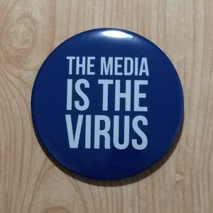 Può includere: Un bottone rotondo blu con testo bianco che recita "THE MEDIA IS THE VIRUS".
