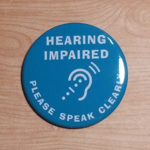 May include: A blue round button with white text that reads "HEARING IMPAIRED" and "PLEASE SPEAK CLEARLY". A white stylized ear with sound waves is in the centre of the button.