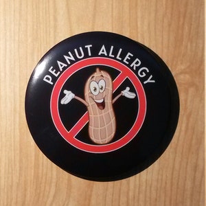 Può includere: Un adesivo rotondo nero e rosso con il testo "Peanut Allergy" e una nocciola di arachidi di cartone animato con una barra rossa.