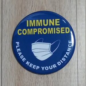 May include: Blue and yellow round sticker with the text "Immune Compromised" and a white face mask graphic. The text "Please Keep Your Distance" is written around the edge of the sticker.