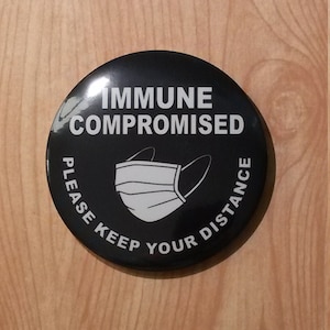 May include: Black and white button with the text "Immune Compromised Please Keep Your Distance" and a white face mask graphic.