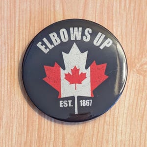 May include: A black button featuring a white and red Canadian flag design. The text "Elbows Up" is above the flag and "Est. 1867" is below the flag.
