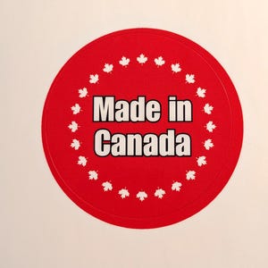 May include: A circular red sticker featuring the text "Made in Canada" in white. The edge of the sticker is decorated with white maple leaf motifs, a symbol of Canada.