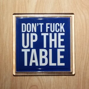Op de afbeelding: Een rechthoekig bord met de tekst "DON'T FUCK UP THE TABLE" in witte letters op een blauwe achtergrond. Het bord is omlijst met een goudkleurige rand en gemonteerd op een licht houten oppervlak.