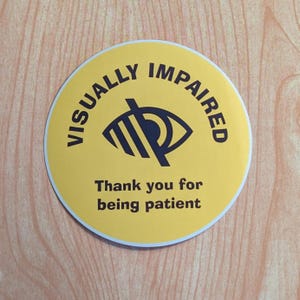 Op de afbeelding: Gele ronde sticker met de tekst "VISUALLY IMPAIRED" in zwarte letters rond een zwart-wit symbool van een doorgestreept oog. De tekst "Thank you for being patient" is in zwart onder het symbool gedrukt.