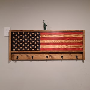 May include: Wooden wall-mounted shelf with an American flag design. The flag features red and natural wood stripes, a dark blue field with stars, and a lower shelf with five black hooks. A small Statue of Liberty figurine sits on top.