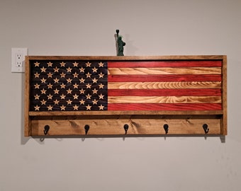 May include: Wooden wall-mounted shelf with an American flag design. The flag features red and natural wood stripes, a dark blue field with stars, and a lower shelf with five black hooks. A small Statue of Liberty figurine sits on top.