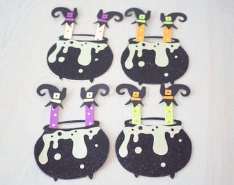 May include: Four black glittery cauldron cutouts with light green bubbling liquid spilling over the rims. Each cauldron features two legs extending upward, wearing patterned socks and pointed black hats with colorful bands in purple, green, and orange.