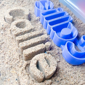 May include: A blue plastic mold and sand create the word "smile". The word is imprinted in the sand, with the blue mold next to it. The sand is a light brown color, and the mold is a bright blue.