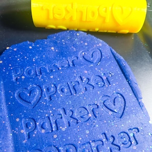 May include: Blue play dough with the name "Parker" imprinted on it three times with a heart symbol after each name. A yellow roller with the word "Parker" and a heart symbol is in the background.