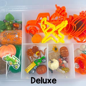 May include: A clear plastic storage container filled with fall-themed craft supplies. Includes orange pumpkin and truck-shaped cookie cutters, a wooden rolling pin, and various decorative elements like pumpkins, leaves, and a toy tractor. The word "Deluxe" is printed on the bottom.