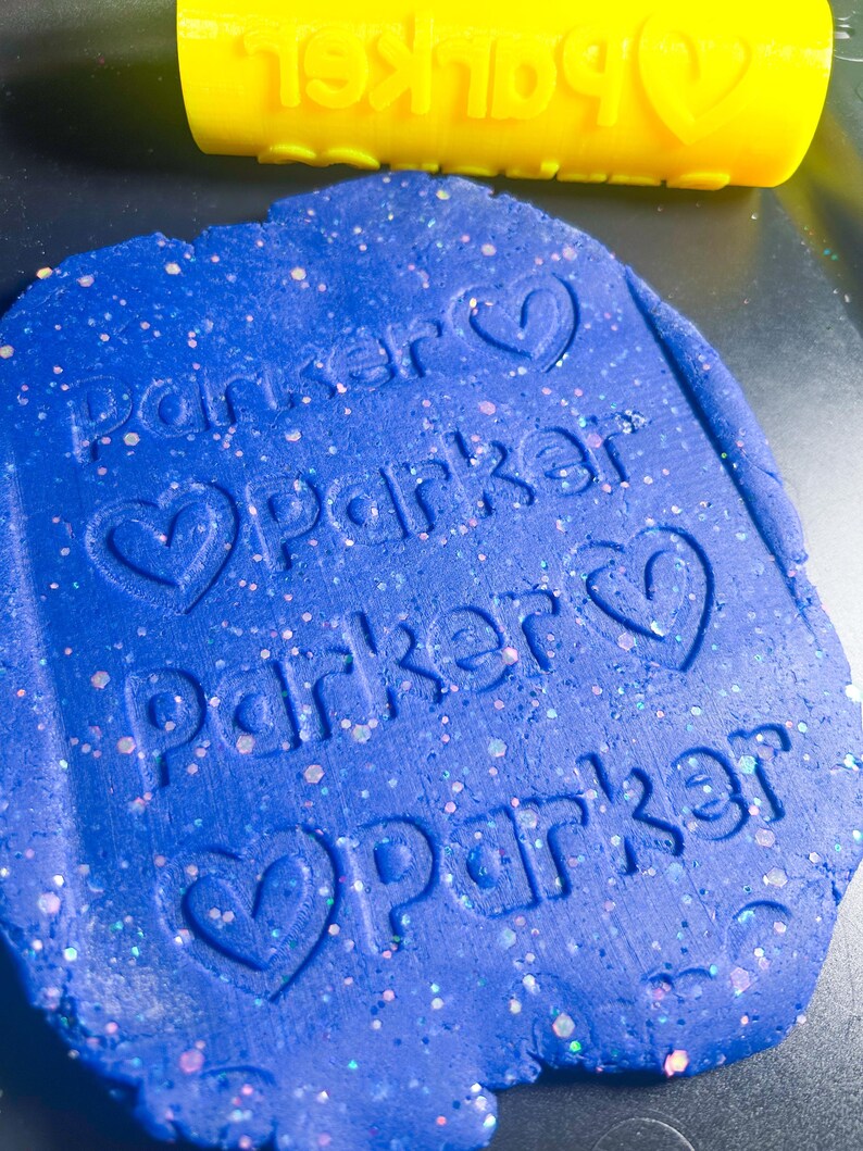 Może przedstawiać: Niebieska masa plastyczna z imieniem "Parker" odciśniętym wielokrotnie z symbolem serca obok każdego imienia. W tle widoczny jest ż&oacute;łty wałek do ciasta z tym samym odciskiem.