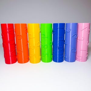 May include: A set of seven cylindrical rollers in a rainbow color sequence of red, orange, yellow, green, blue, purple, and pink. Each roller features raised, embossed letters and numbers on its surface, arranged in a vertical orientation.