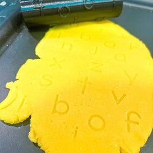 May include: A yellow playdough with stamped letters and a black roller stamp. The playdough has the letters "b, i, o, f, t, v, j, n, z, s, a" stamped into it. The roller stamp is black with a blue stripe.