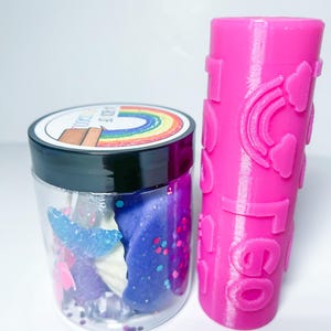 May include: A clear jar filled with purple and blue slime, glitter, and a blue mermaid tail charm. Next to the jar is a pink roller with a rainbow and heart design.