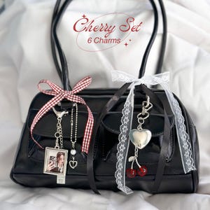 May include: Black handbag with two charms. One charm features a photo in a silver frame, the other has a silver heart and red cherry charms. The bag has black handles and is decorated with red and white gingham ribbon and white lace.