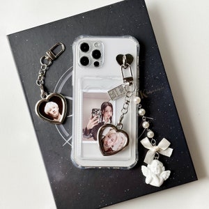 Może przedstawiać: Przezroczyste etui na telefon ze zdjęciem i zawieszkami w kształcie serca. Etui ma zdjęcie osoby i zawieszkę w kształcie serca ze zdjęciem. Do etui dołączony jest urok perłowy i anioła. Etui jest na ciemnym tle.