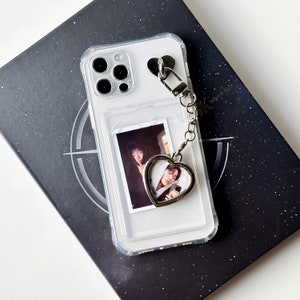 Pode incluir: Uma capa transparente para telefone com um amuleto em forma de coração preso a uma corrente. A capa tem um suporte para fotos integrado com uma foto de duas pessoas. A capa está sobre um fundo preto.