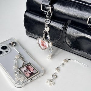Può includere: Due ciondoli per telefono con accenti di perle e cristalli. Un ciondolo presenta una cornice per foto a forma di cuore con una foto di una persona. L'altro ciondolo ha una cornice per foto rettangolare con una foto di una persona. Entrambi i ciondoli hanno un fiocco bianco.
