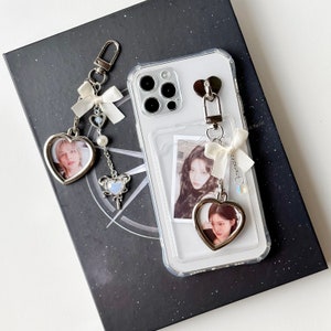 May include: A clear phone case with a white and silver charm attached. The charm has a heart-shaped photo frame with a picture of a person inside. The phone case has a built-in pocket for a photo card. The phone case is on a black surface.