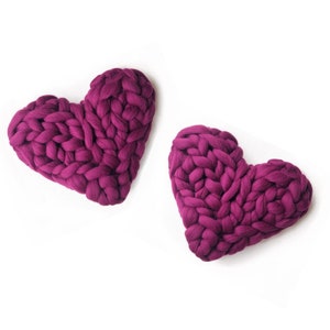 May include: Two large, knitted heart-shaped cushions in a deep purple colour. The cushions are made of chunky yarn and have a soft, textured surface.