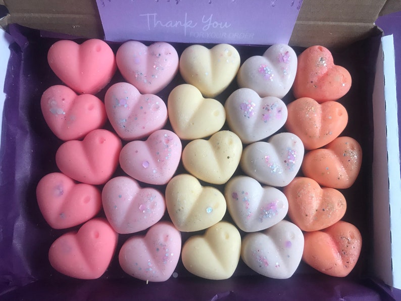 May include: A box filled with 27 heart-shaped wax melts in three colors: pink, yellow, and orange. Some of the melts have glitter on them.