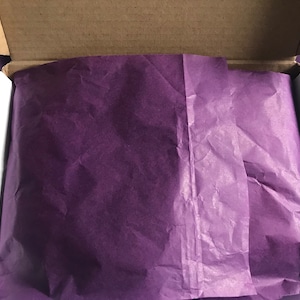 May include: An open cardboard box lined with purple tissue paper. The tissue paper is crumpled and has a soft, delicate texture.