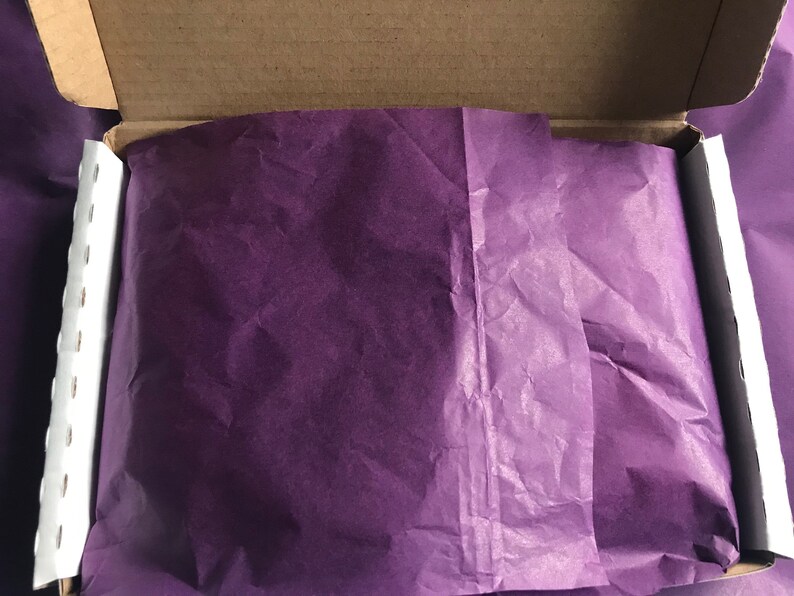 May include: An open cardboard box lined with purple tissue paper. The tissue paper is crumpled and has a soft, delicate texture.