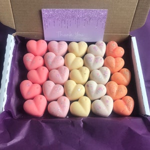 May include: A box filled with 24 heart-shaped wax melts in shades of pink, yellow, and orange. The wax melts are arranged in rows, with the pink melts on the bottom, the yellow melts in the middle, and the orange melts on top. The box is lined with purple tissue paper and has a "Thank You" card on top.