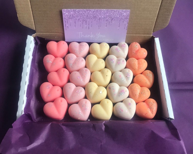 May include: A box filled with 24 heart-shaped wax melts in shades of pink, yellow, and orange. The wax melts are arranged in rows, with the pink melts on the bottom, the yellow melts in the middle, and the orange melts on top. The box is lined with purple tissue paper and has a "Thank You" card on top.