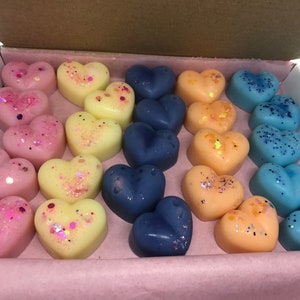 May include: A box of heart-shaped wax melts in pink, yellow, and blue colors. The melts are decorated with glitter and are arranged in rows.