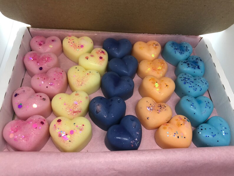 May include: A box of heart-shaped wax melts in pink, yellow, and blue colors. The melts are decorated with glitter and are arranged in rows.