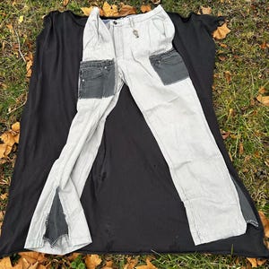 Op de afbeelding: Lichtgrijze broek met zwarte denim zakpatches en zijsplitten. De broek heeft een sleutelhanger aan de riemlus en ligt op een zwarte stof. De achtergrond is een bed van gevallen bladeren en gras.