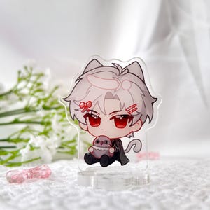 May include: A clear acrylic keychain featuring a cartoon character with white hair, red eyes, and a pink bow. The character is holding a small, gray, plush animal. The keychain has a cat ear design.