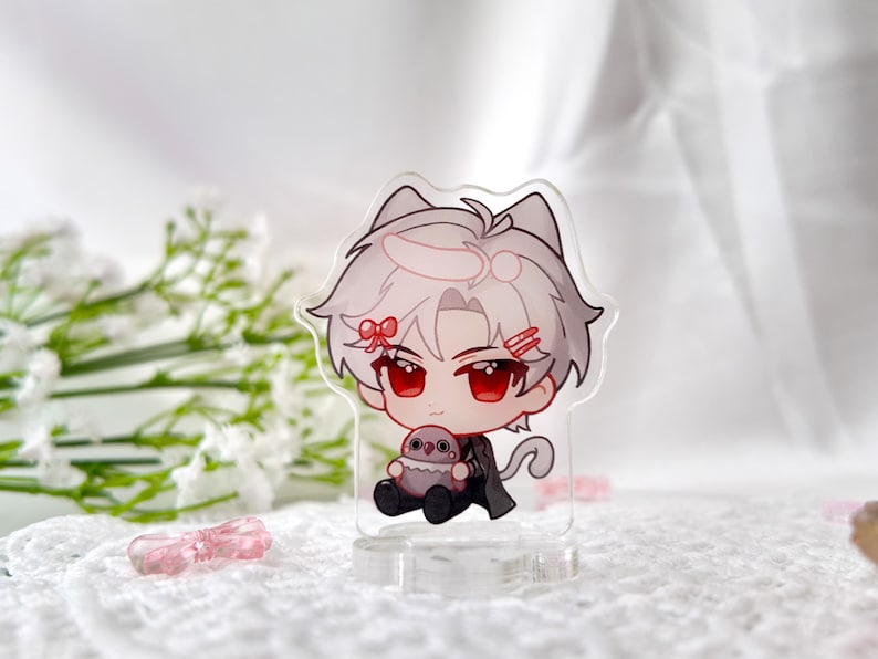 May include: A clear acrylic keychain featuring a cartoon character with white hair, red eyes, and a pink bow. The character is holding a small, gray, plush animal. The keychain has a cat ear design.
