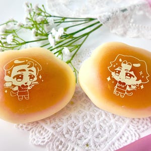 Könnte beinhalten: Zwei goldbraune, herzförmige Gebäckstücke mit Cartoon-Illustrationen. Jedes Gebäckstück hat ein anderes Charakterdesign. Das Gebäck liegt auf einer weißen Spitzendecke mit weißen Blumen und rosa Schleifen.