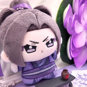 Peut inclure: Une poupée en peluche aux cheveux gris coiffés en chignon, vêtue d'une tenue violette avec des accents sombres. La poupée présente des traits brodés, dont des yeux foncés et une expression sévère. Un petit pot marron est posé sur une table noire.