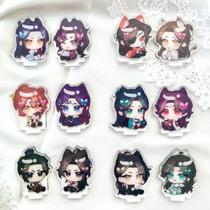 May include: A collection of acrylic anime character standees. Each standee features a chibi-style illustration of a character with detailed hair and clothing. The characters have various hair colors and are decorated with bows and other accessories. The standees are on a white surface.