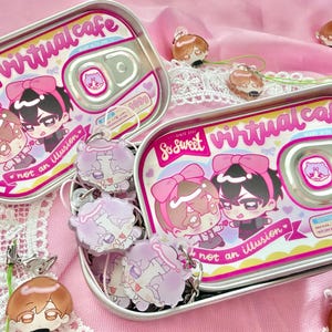 May include: Two tin containers with pink and white labels that read "So Sweet virtual cafe" and "not an illusion" with cartoon characters. Several matching keychains are also visible, along with other keychains.