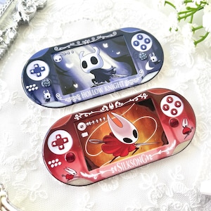 May include: Two handheld gaming console cases. One is navy blue with a Hollow Knight design, the other is red with a Silksong design. Both have game controller button layouts and decorative borders. The cases are on a white lace surface.