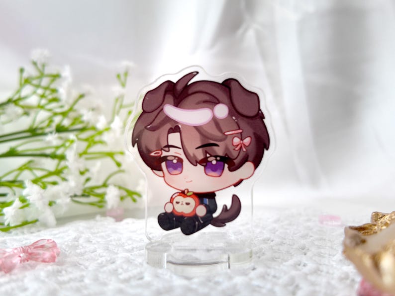 May include: A clear acrylic stand with a cartoon character holding an apple. The character has brown hair, purple eyes, and is wearing a black and white outfit. The character has dog ears and a pink bow in their hair.