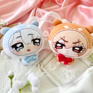 以下が含まれることがあります： 2つのぬいぐるみキャラクターキーホルダー。1つは水色で青い蝶ネクタイ、もう1つはオレンジ色で赤いリボンが付いています。どちらも大きな目と漫画の顔をしています。白いレースの表面に花とピンクのサテンの背景があります。