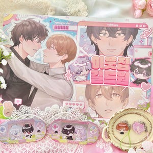 May include: An illustration with anime-style characters, focusing on romance. The image includes a print with Korean text, two handheld game-shaped stickers, and a decorative tray with charms. The colour scheme is soft, with pinks and pastels.