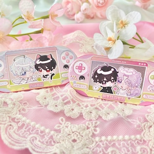 May include: Two pink handheld game consoles with cartoon character graphics. One features a black-haired character with cat ears and a white-haired character. The other has two characters with black and purple hair. The text "illusion.exe" is visible on the screens.