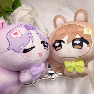May include: Two plush toys featuring cartoon-style faces. The left plush is light purple with a heart speech bubble and a purple bow. The right plush is tan with brown ears, a question mark speech bubble, and a bright green bow. Both have large eyes.