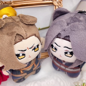 Puede incluir: Dos muñecos de peluche con cabello suave marrón y gris, con expresiones faciales bordadas y ojos amarillos. Cada muñeco viste un atuendo detallado con patrones en azul oscuro y marrón, y uno tiene un accesorio de pelo amarillo brillante.