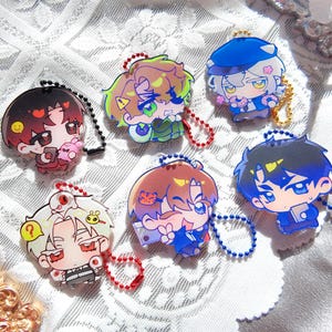 Puede incluir: Una colección de cinco dijes de acrílico con personajes de anime estilo chibi, con cabello colorido y rostros expresivos. Cada dije está sujeto a una pequeña cadena de bolas de metal en varios colores, incluyendo negro, rojo, azul y dorado.